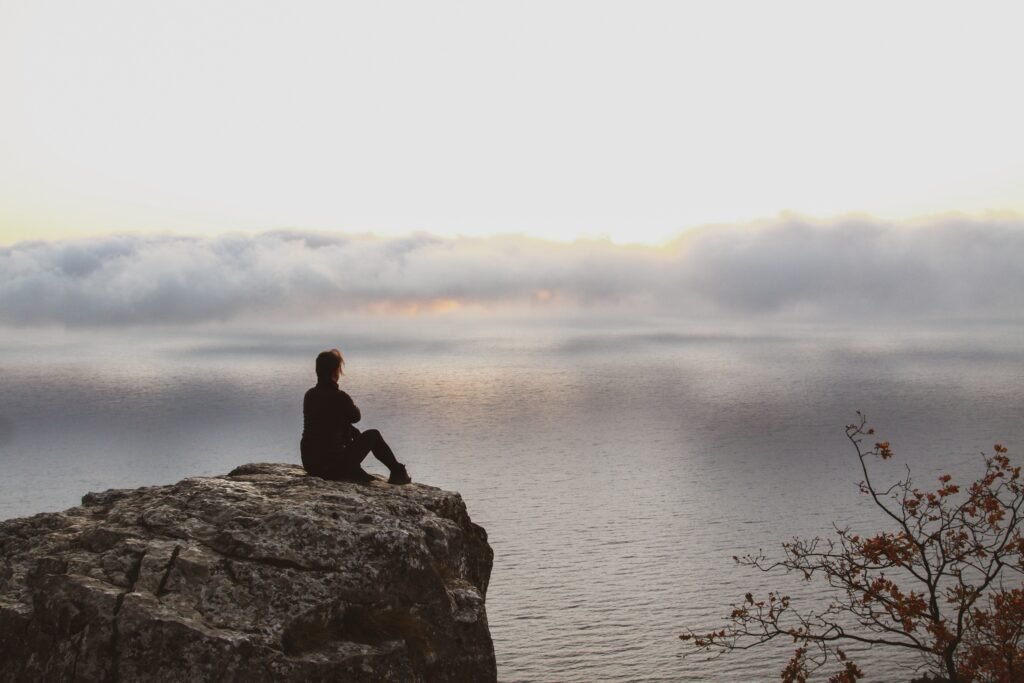 A person sitting alone on a rocky cliff, looking out over calm water and misty clouds during sunrise.