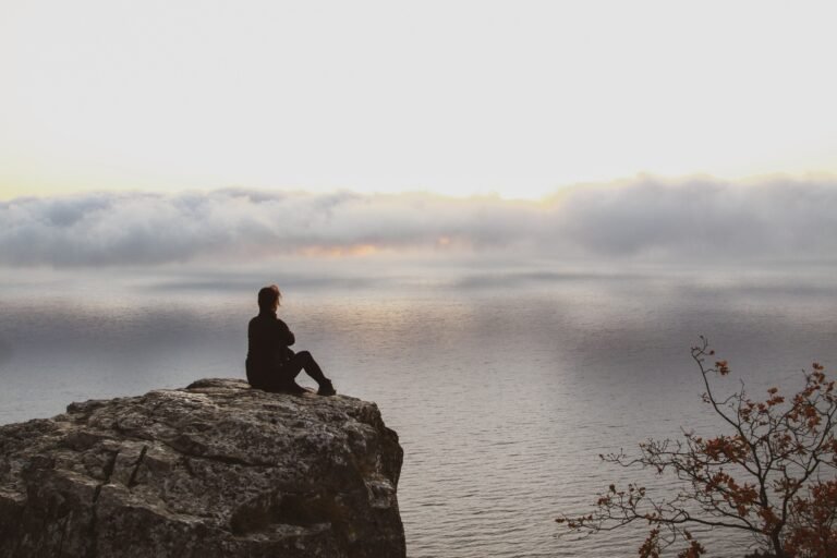 A person sitting alone on a rocky cliff, looking out over calm water and misty clouds during sunrise.