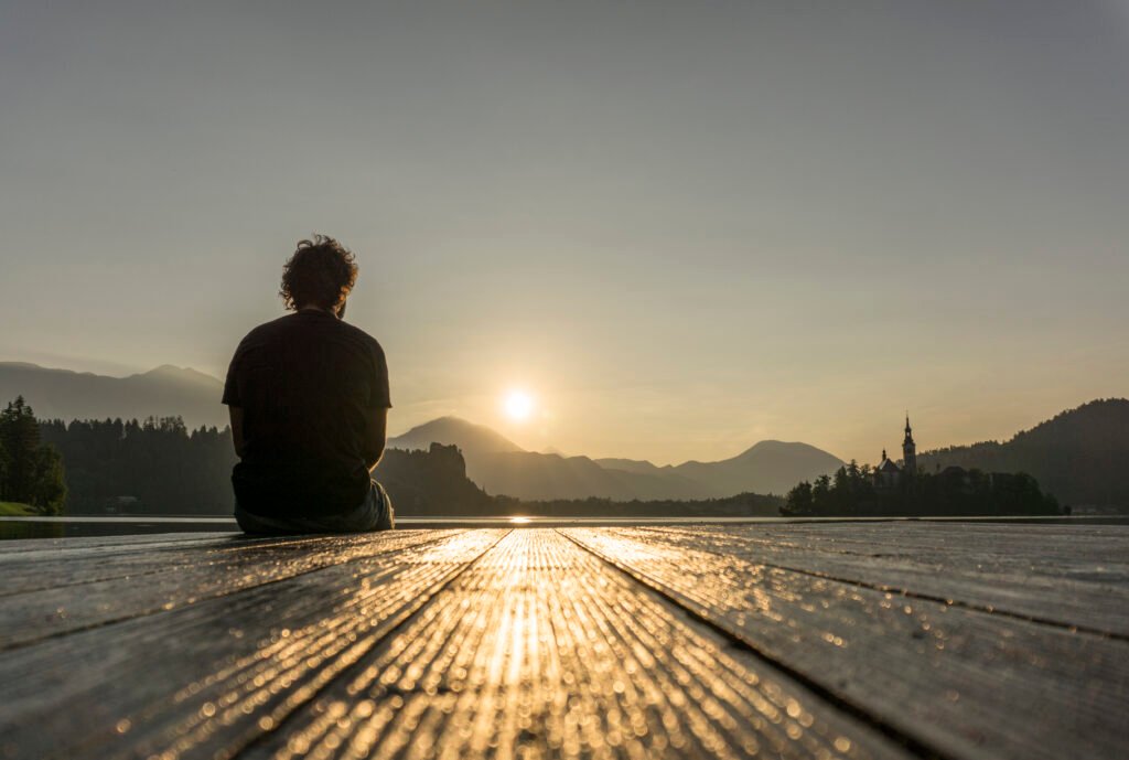 A man sitting alone on a wooden pier, watching the sunrise over a calm lake with mountains in the background.