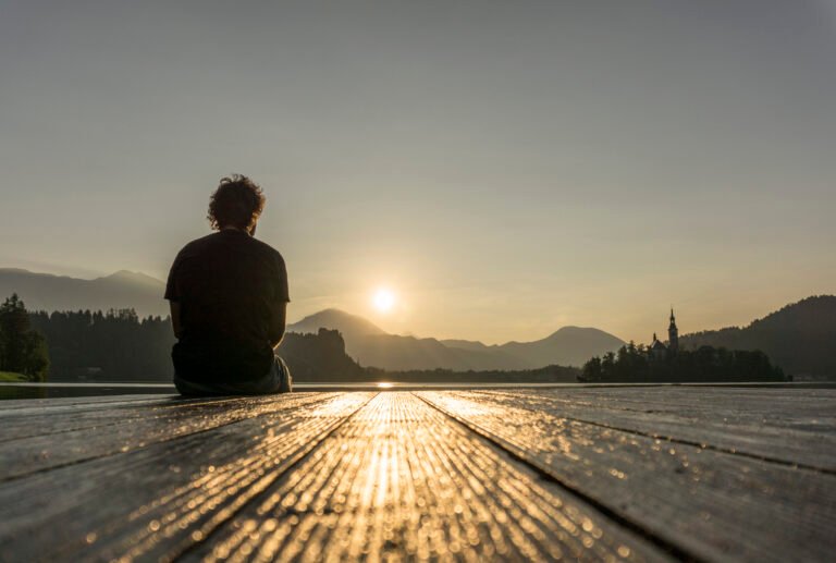 A man sitting alone on a wooden pier, watching the sunrise over a calm lake with mountains in the background.
