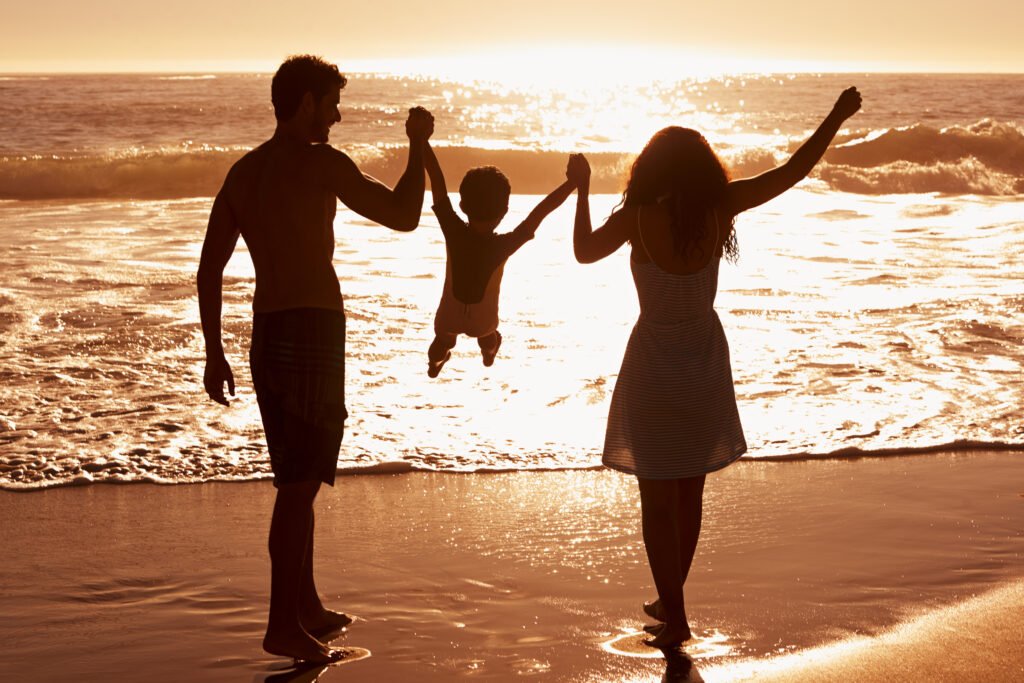 Parents holding their child’s hands and lifting them in the air while standing on a beach at sunset.