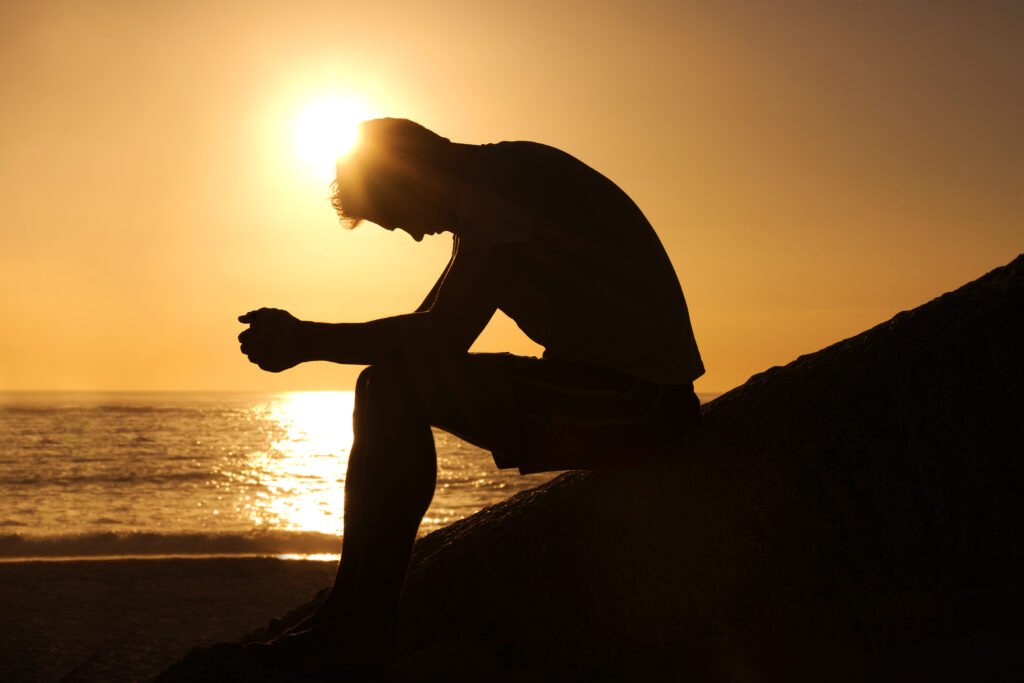 A silhouette of a man sitting on a rock by the ocean at sunset, with his head bowed in deep thought.
