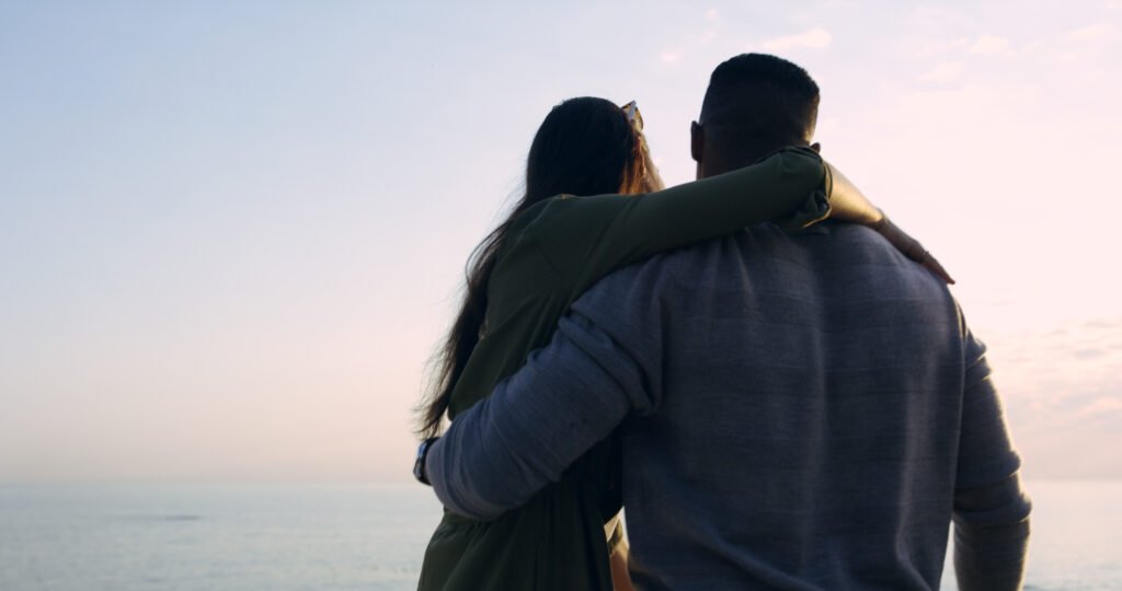 A couple standing close together with arms around each other, looking at the ocean during a peaceful sunset.