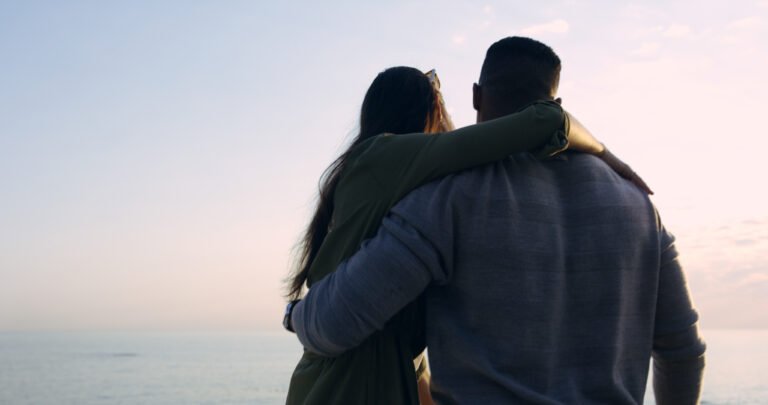 A couple standing close together with arms around each other, looking at the ocean during a peaceful sunset.