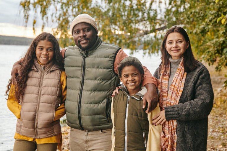 A happy family of four standing together outdoors near a lake during autumn, smiling and dressed in warm clothing.